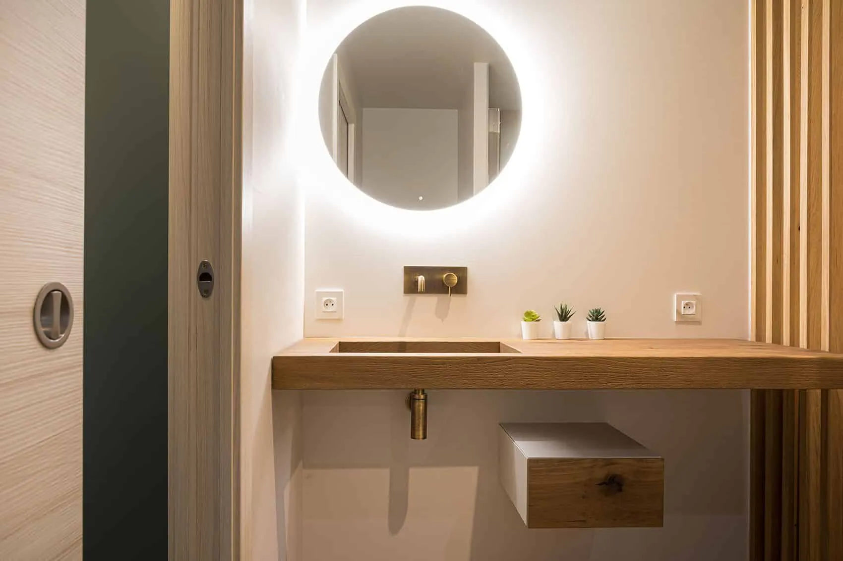 Salle de douche de la suite parentale : miroir rond rétroéclairé, plan vasque en bois et robinetterie murale dorée