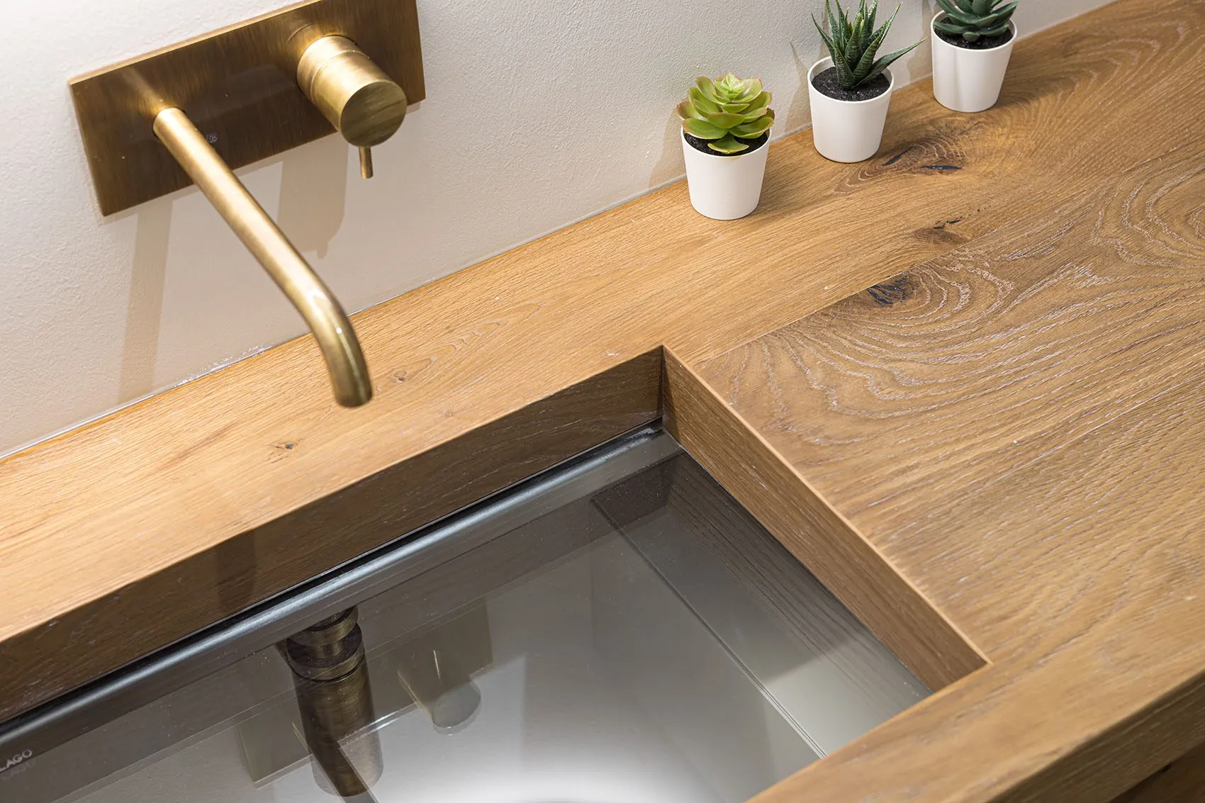 Vasque Lago à fond de verre encastrée dans un plan en bois, robinet mural doré et petites plantes grasses