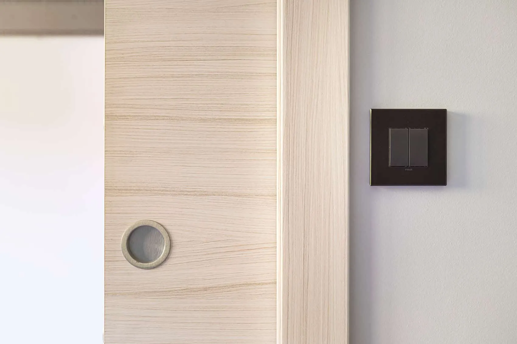 Porte coulissante effet bois clair à poignée cuvette ronde et interrupteur noir sur mur blanc
