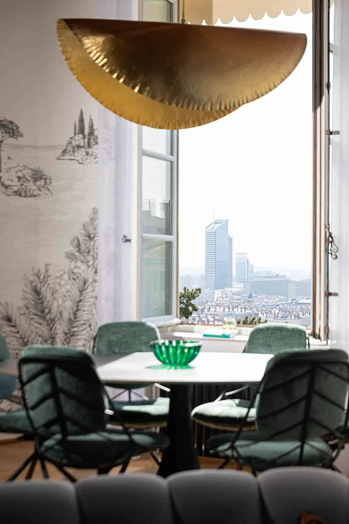 Salle à manger face à la fenêtre ouverte sur les toits de Lyon, chaises en velours vert et papier peint Cypres