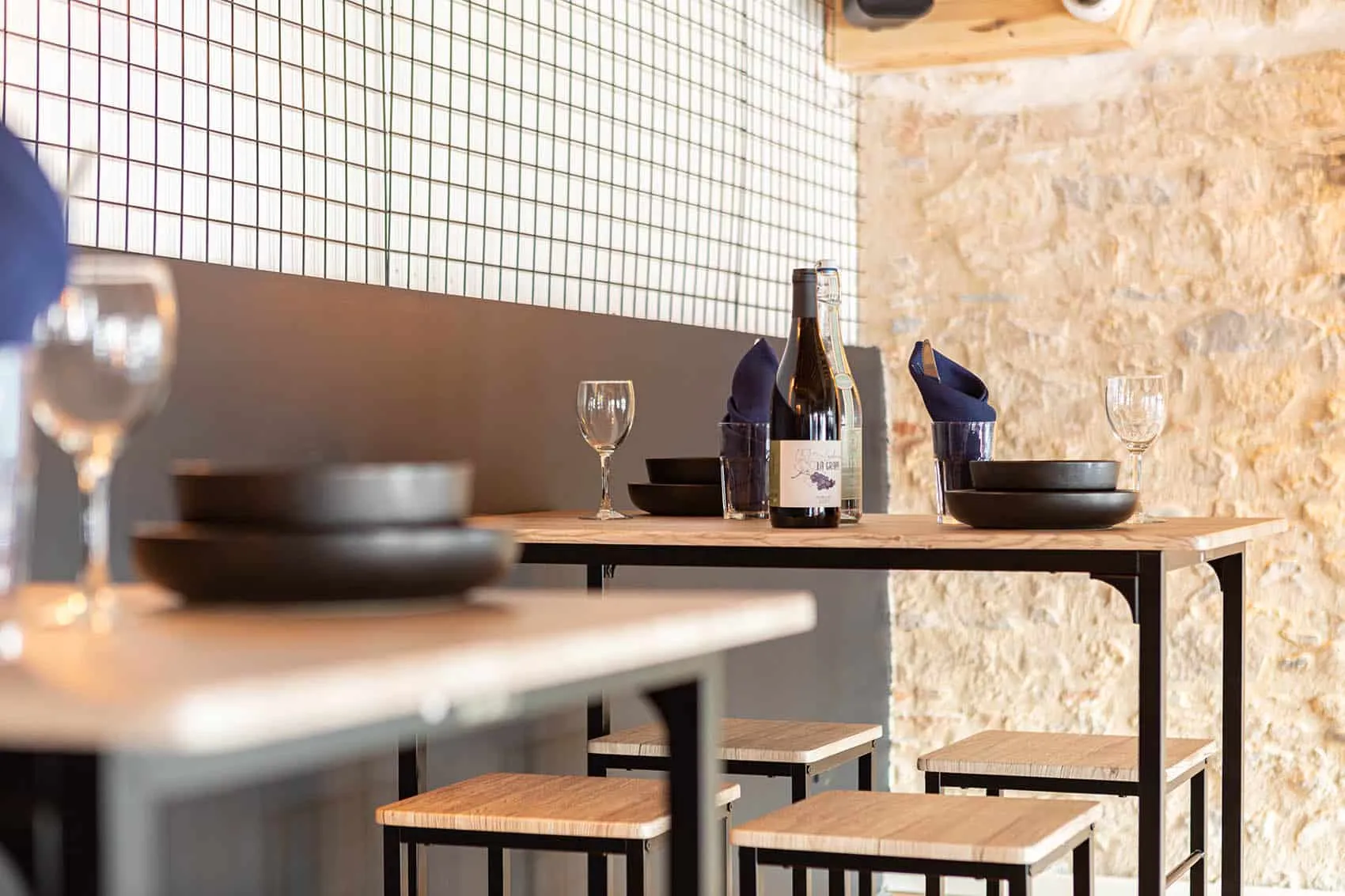Tables hautes en bois et métal noir avec tabourets assortis, mur en pierre et carrelage blanc quadrillé