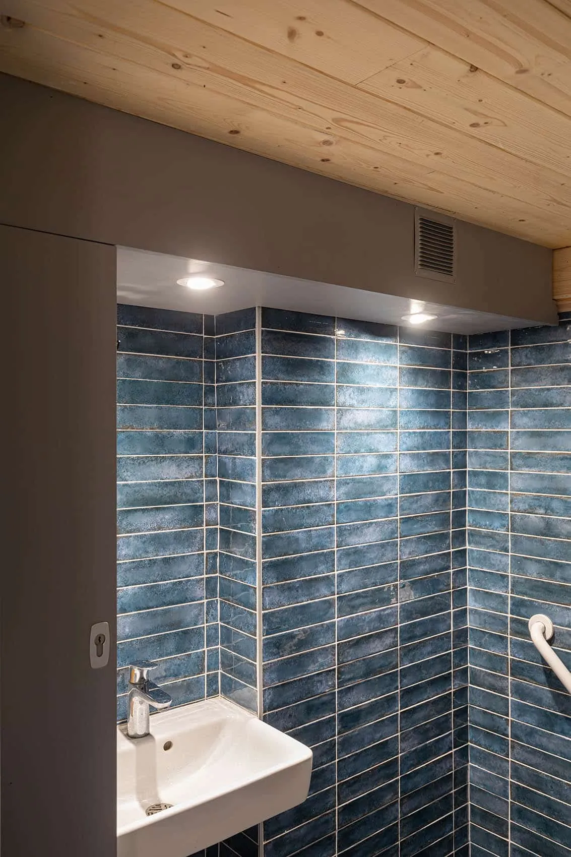 Sanitaires accessibles PMR du restaurant : faïence bleue en longs carreaux, lavabo blanc, barre d’appui et plafond en lambris