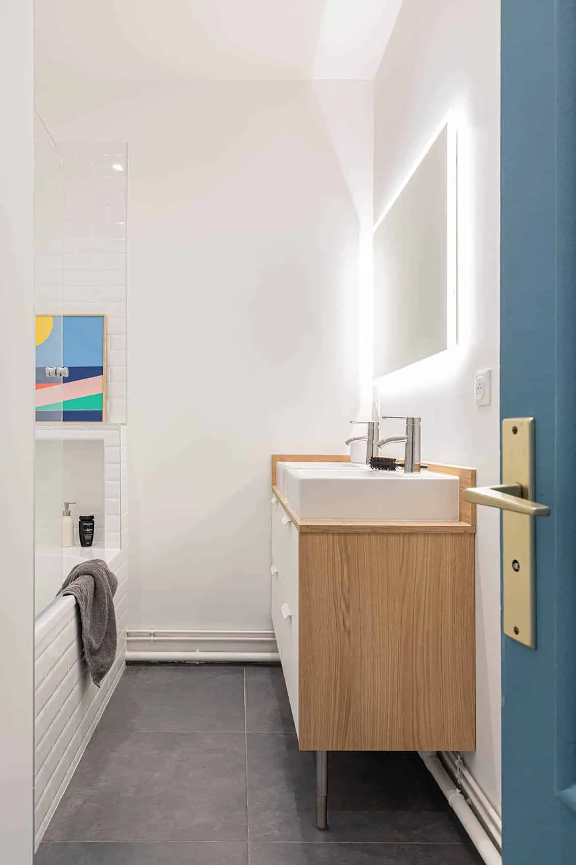 Salle de bain : meuble vasque en bois clair, miroir rétroéclairé et affiche colorée au-dessus de la baignoire