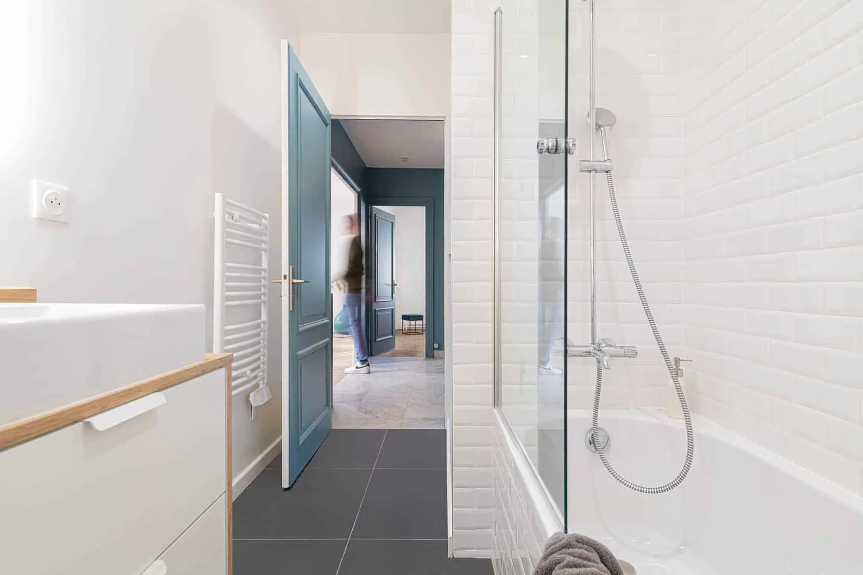 Douche au-dessus de la baignoire, sèche-serviettes blanc et porte bleue ouvrant sur le couloir