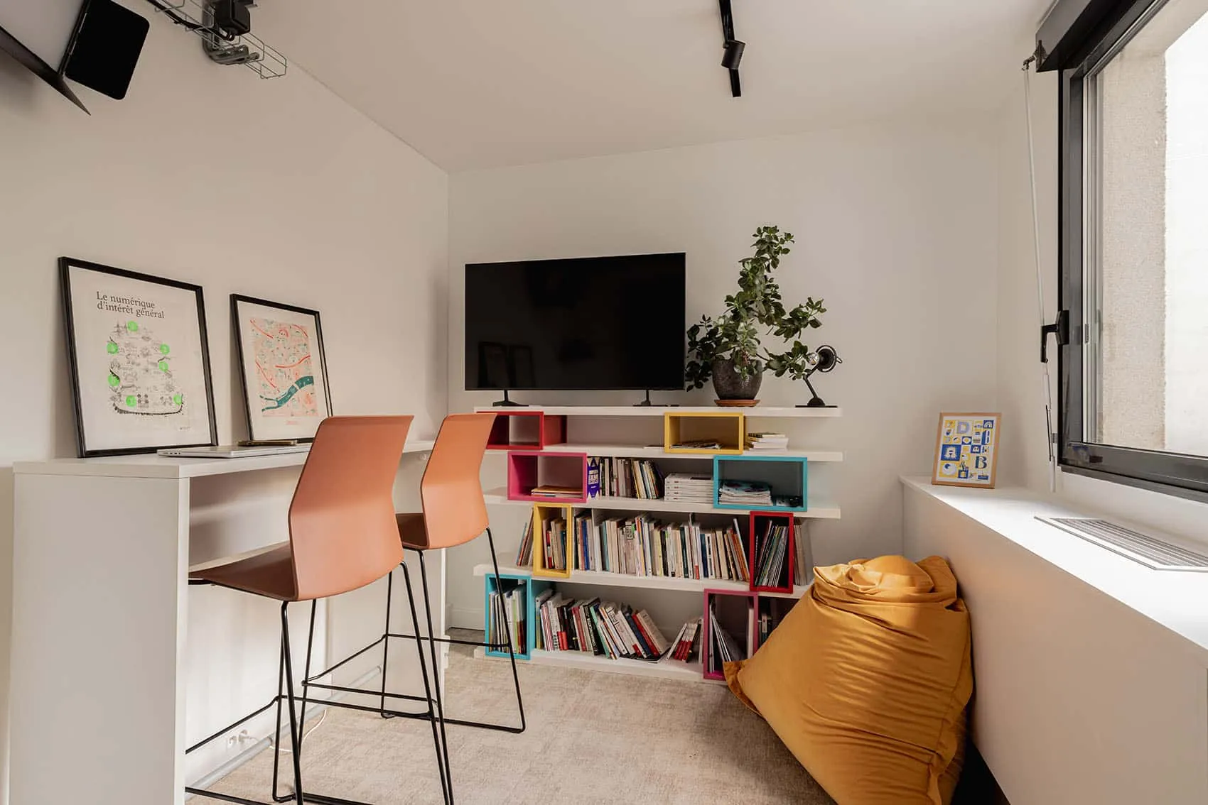 Salle de pause avec bibliothèque colorée, tabourets orange et pouf jaune, design de bureaux