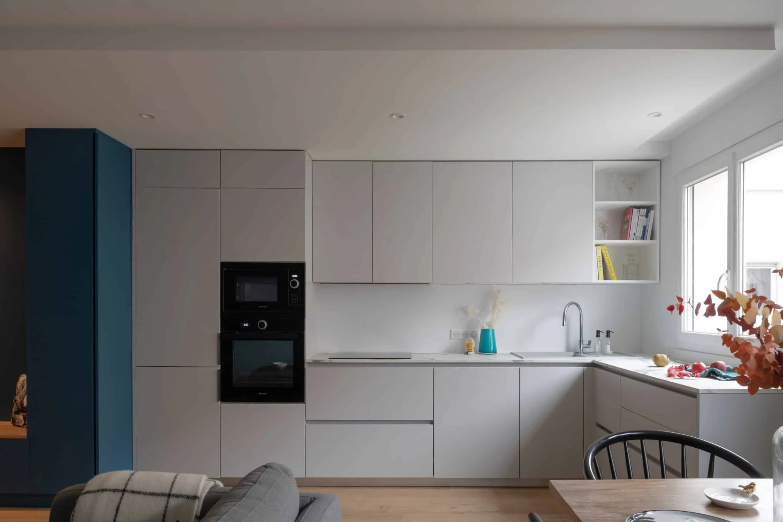 Cuisine ouverte sur le séjour, façades gris clair, fours encastrés et colonne bleue, rénovation d’appartement à Lyon