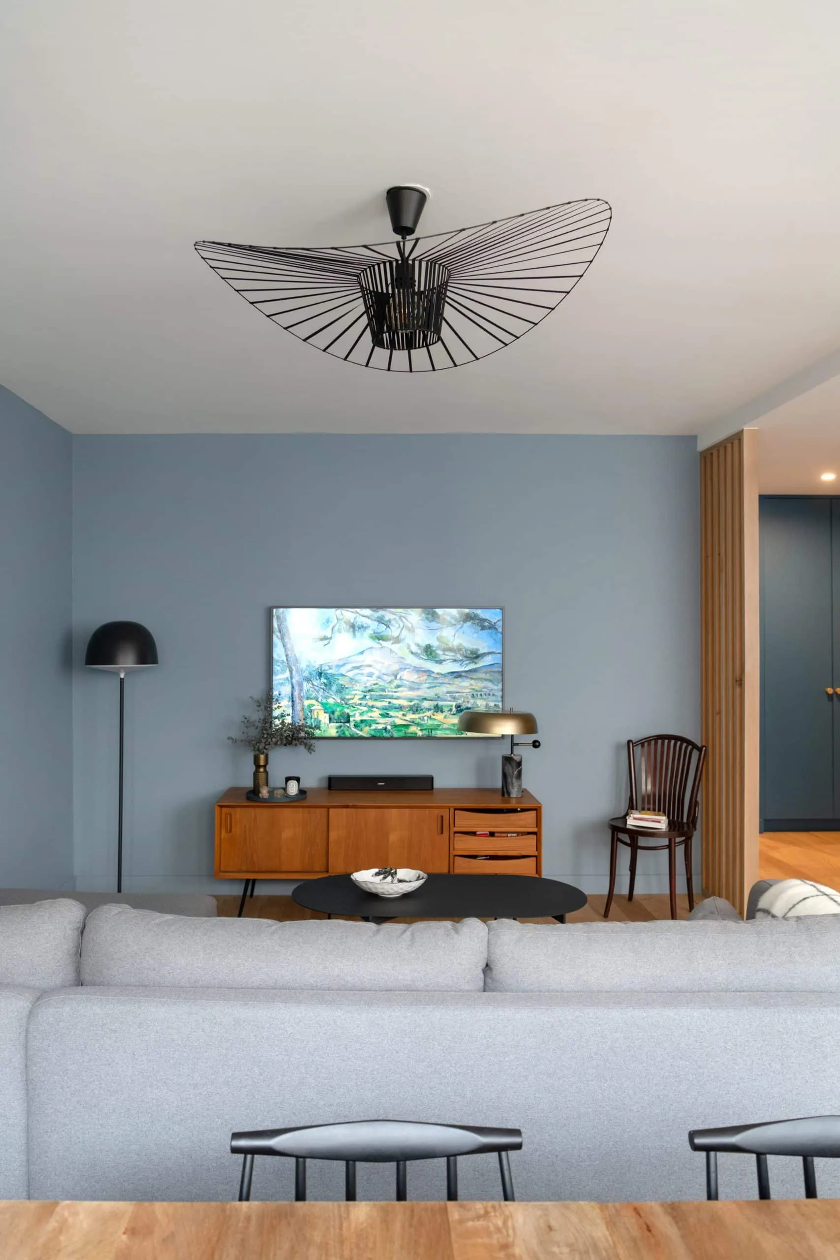 Coin télévision sur mur bleu-gris avec enfilade en bois vintage, lampadaire noir et chaise bistrot