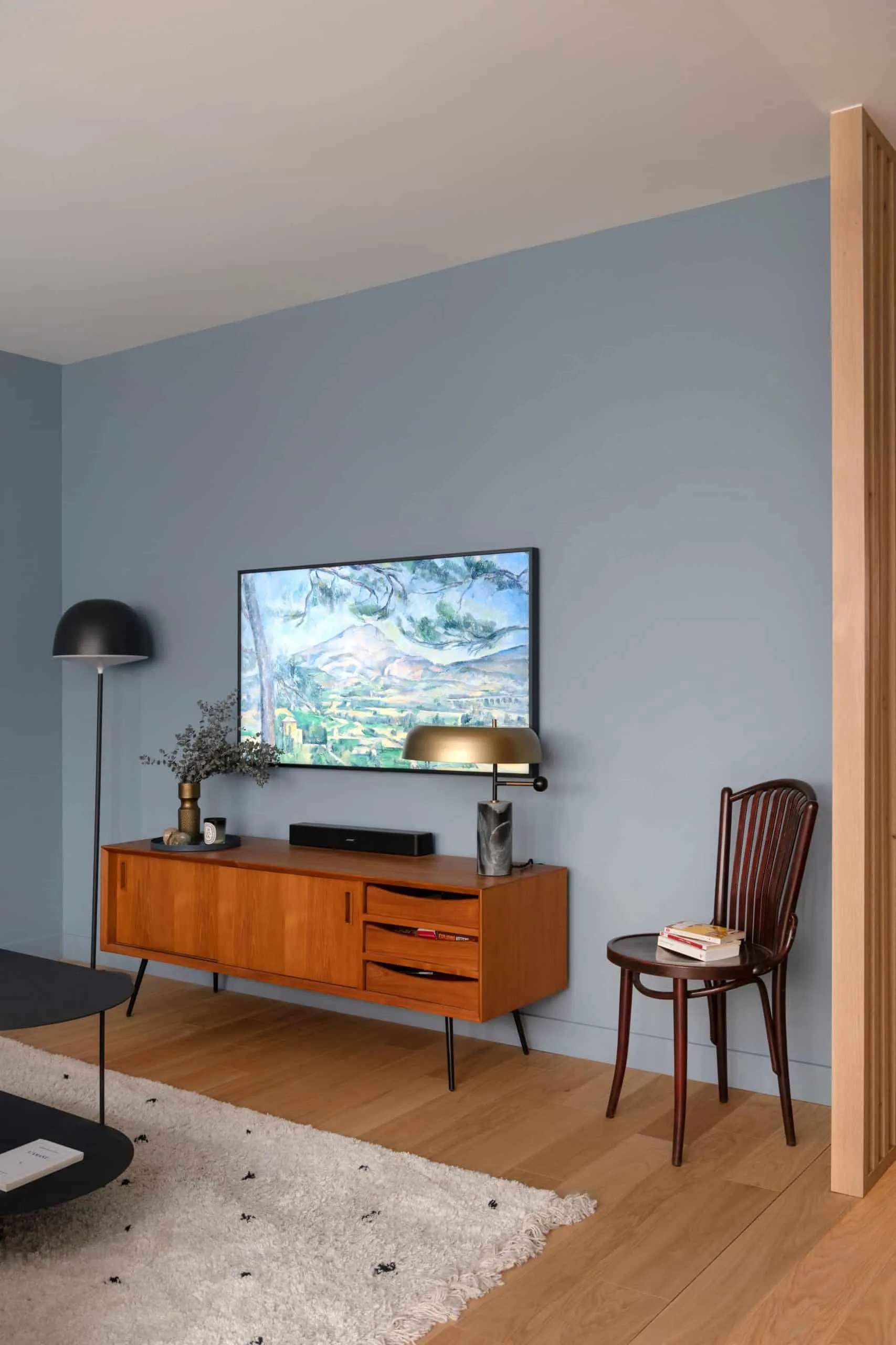 Enfilade vintage en bois, lampe dorée et écran affichant un paysage peint sur le mur bleu-gris du salon