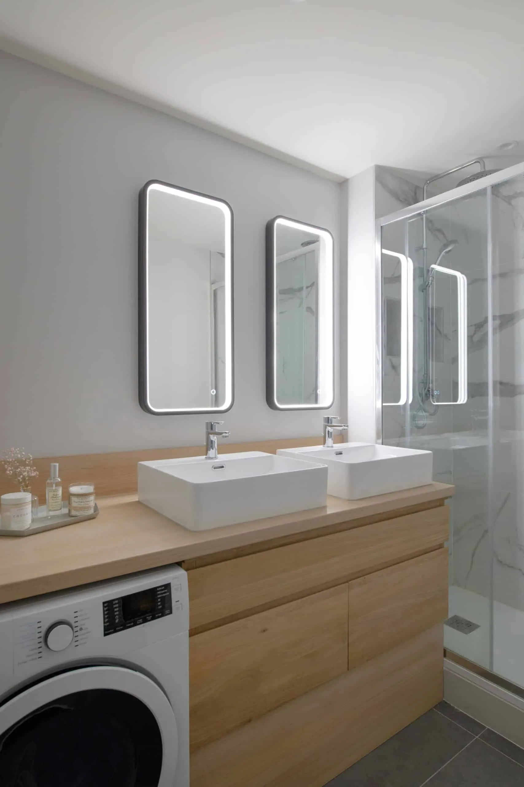 Salle de bains avec double vasque sur plan en bois clair, miroirs rétroéclairés et douche vitrée