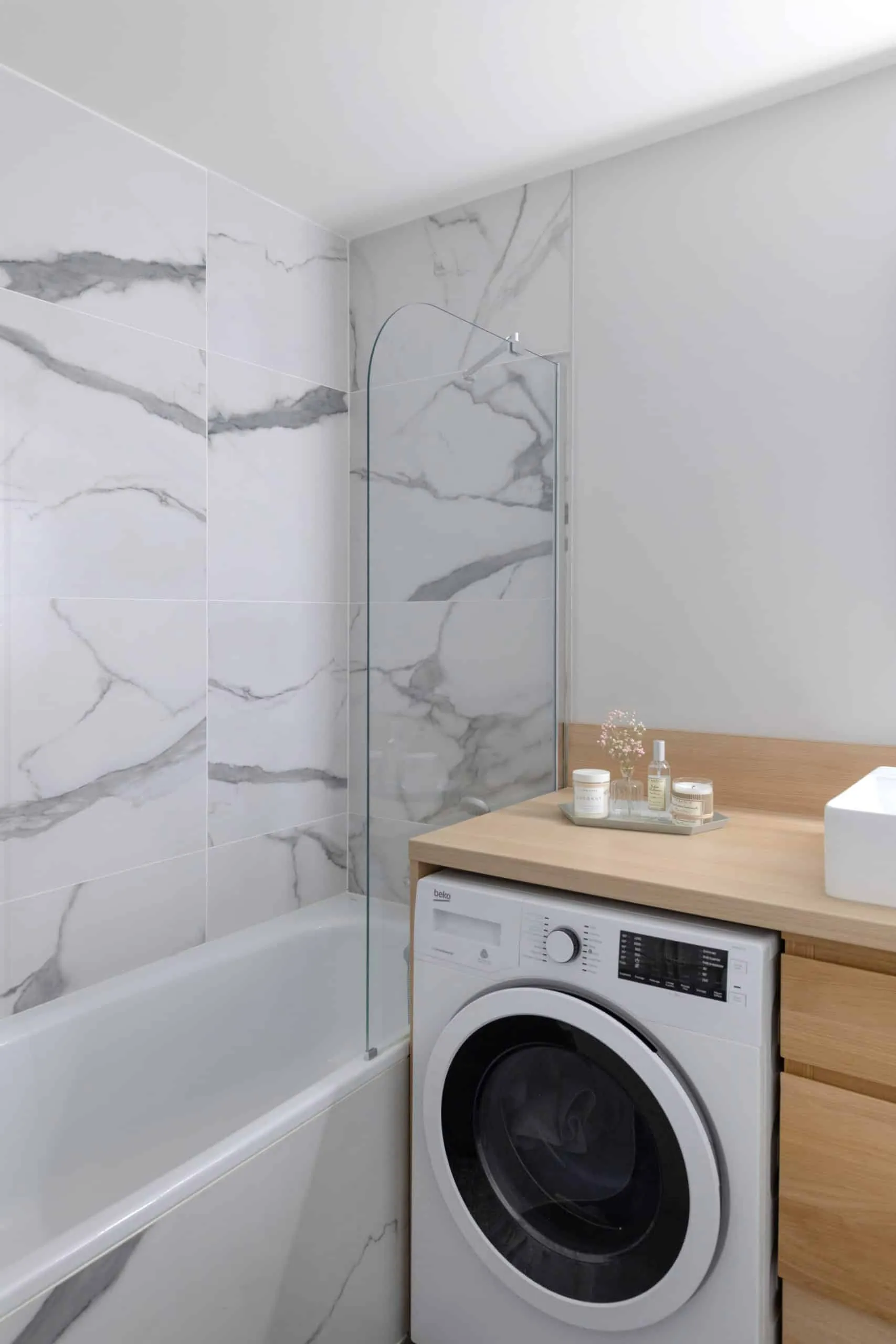 Salle de bains au carrelage effet marbre blanc, plan en bois clair et lave-linge intégré sous le plan
