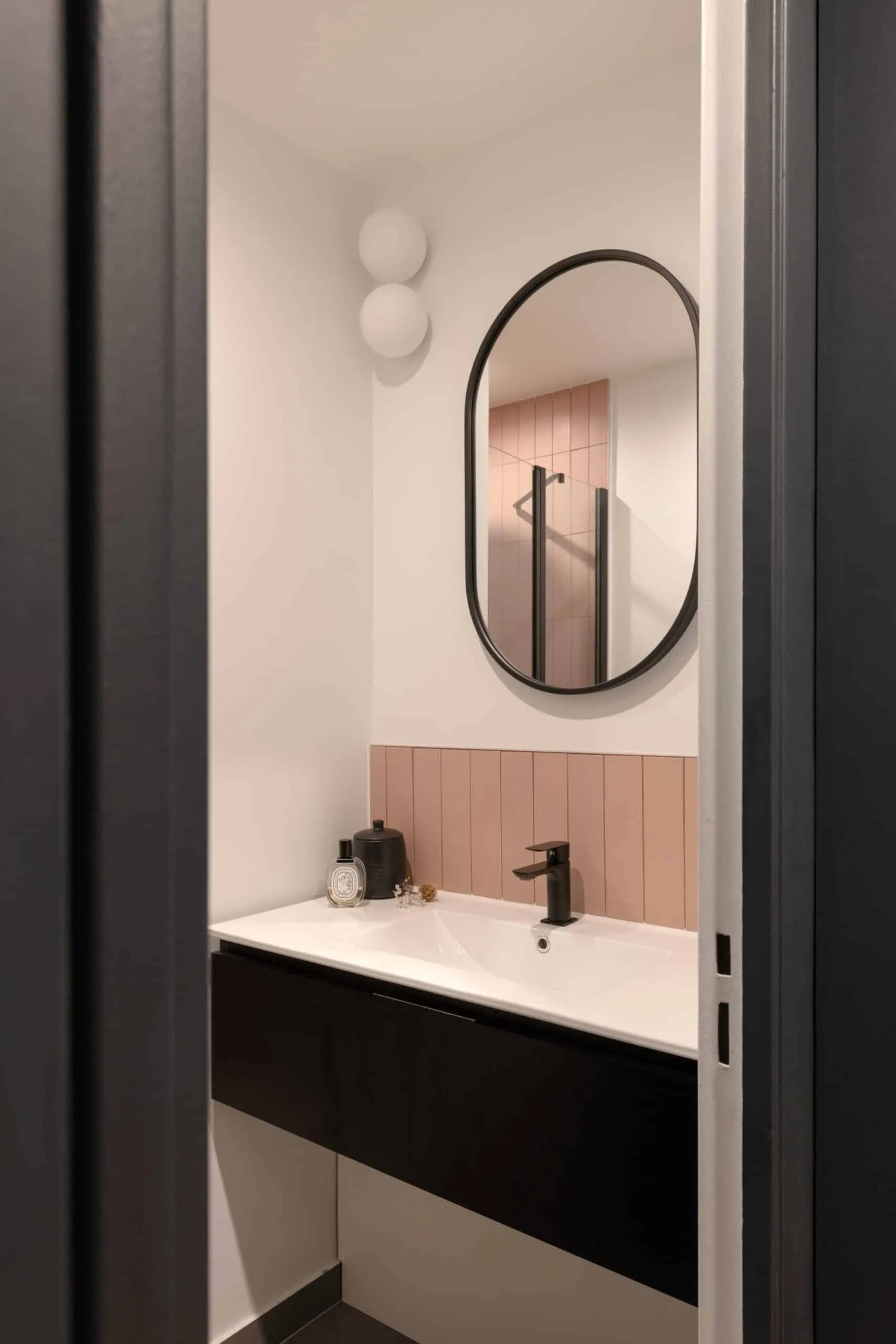 Salle d’eau aux carreaux verticaux vieux rose, vasque blanche, robinet noir et miroir ovale cerclé de noir