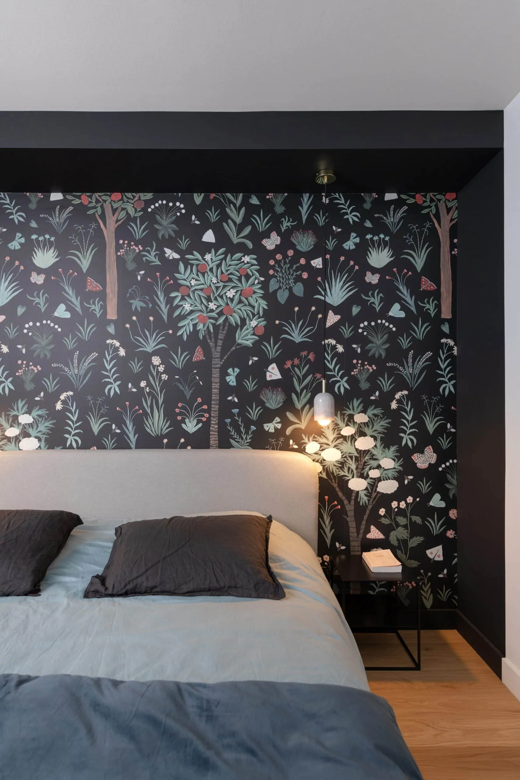 Chambre au papier peint végétal sur fond noir, tête de lit blanche et suspension blanche allumée