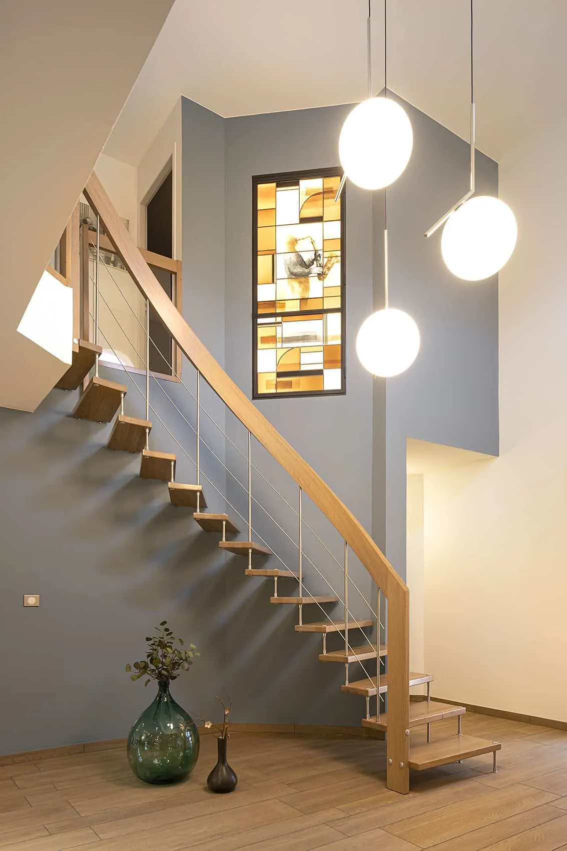 Cage d’escalier au mur bleu doux, vitrail coloré sur mesure et suspensions globes chromées, architecte d’intérieur à Lyon