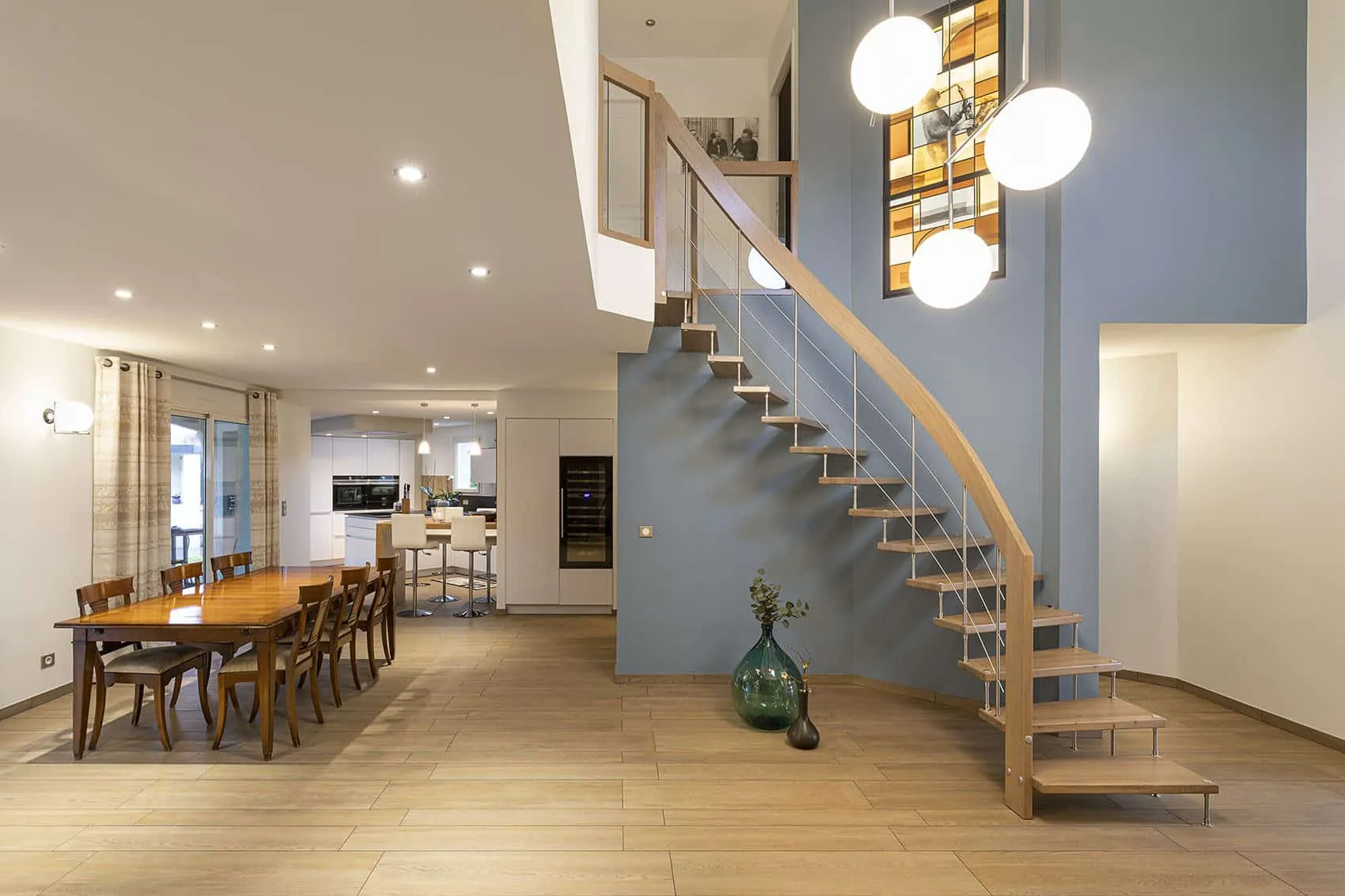 Vue d’ensemble de la pièce de vie rénovée : salle à manger en bois, escalier suspendu et vitrail Art déco