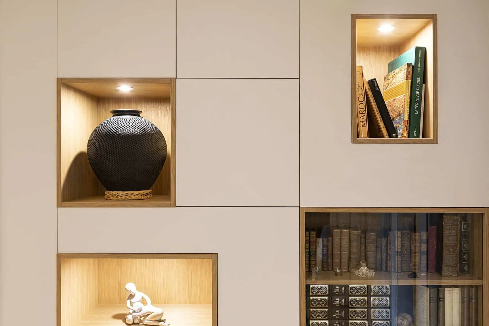 Niches éclairées en bois clair dans la bibliothèque blanche : vase noir, statuette et livres anciens sous vitrine