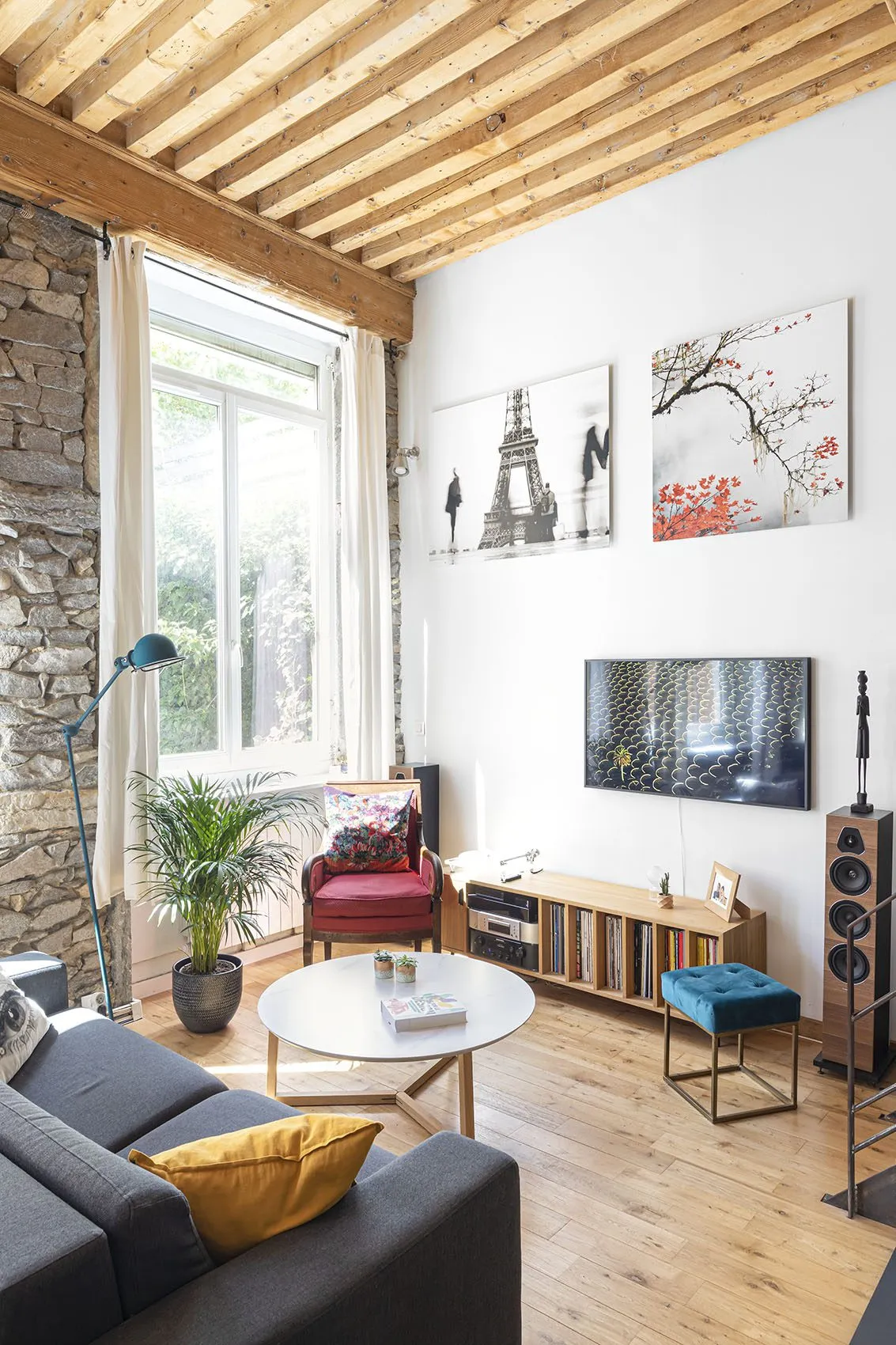 Coin salon du loft canut, entre mur en pierre apparente et grandes fenêtres : fauteuil rouge, meuble vinyles et parquet en bois