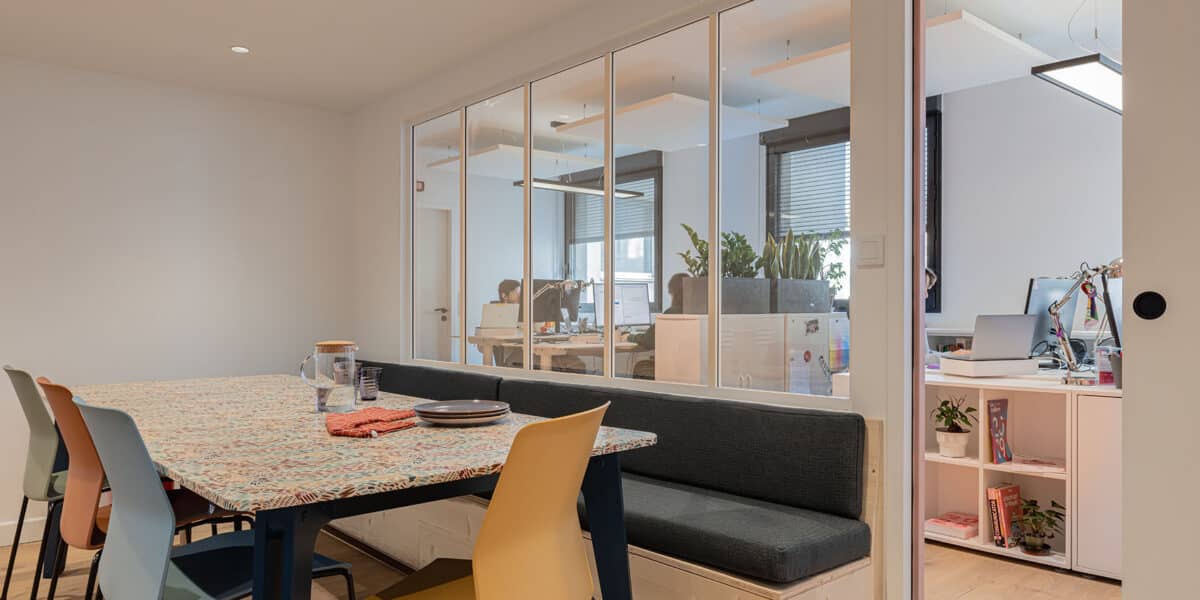 Salle de pause contemporaine avec banquette sur mesure et bureaux ouverts séparés par des verrières, réalisée par une agence d'architecture d'intérieur à Lyon.