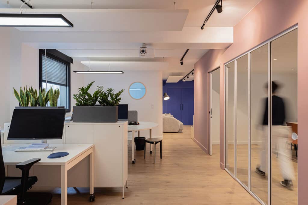 Aménagement de bureaux en open space avec cloisons vitrées, murs roses et bleu nuit, mobilier blanc et plantes vertes