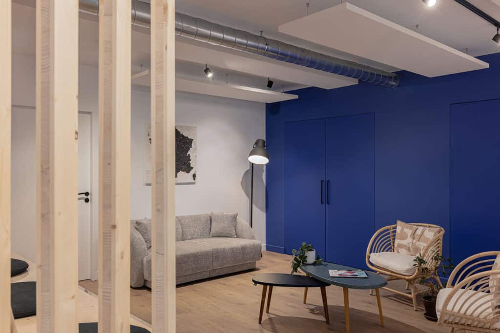 Espace de détente aménagé dans des bureaux à Lyon : cloison bois, canapé gris, fauteuils en rotin et mur bleu Klein, réalisé par Picture Design